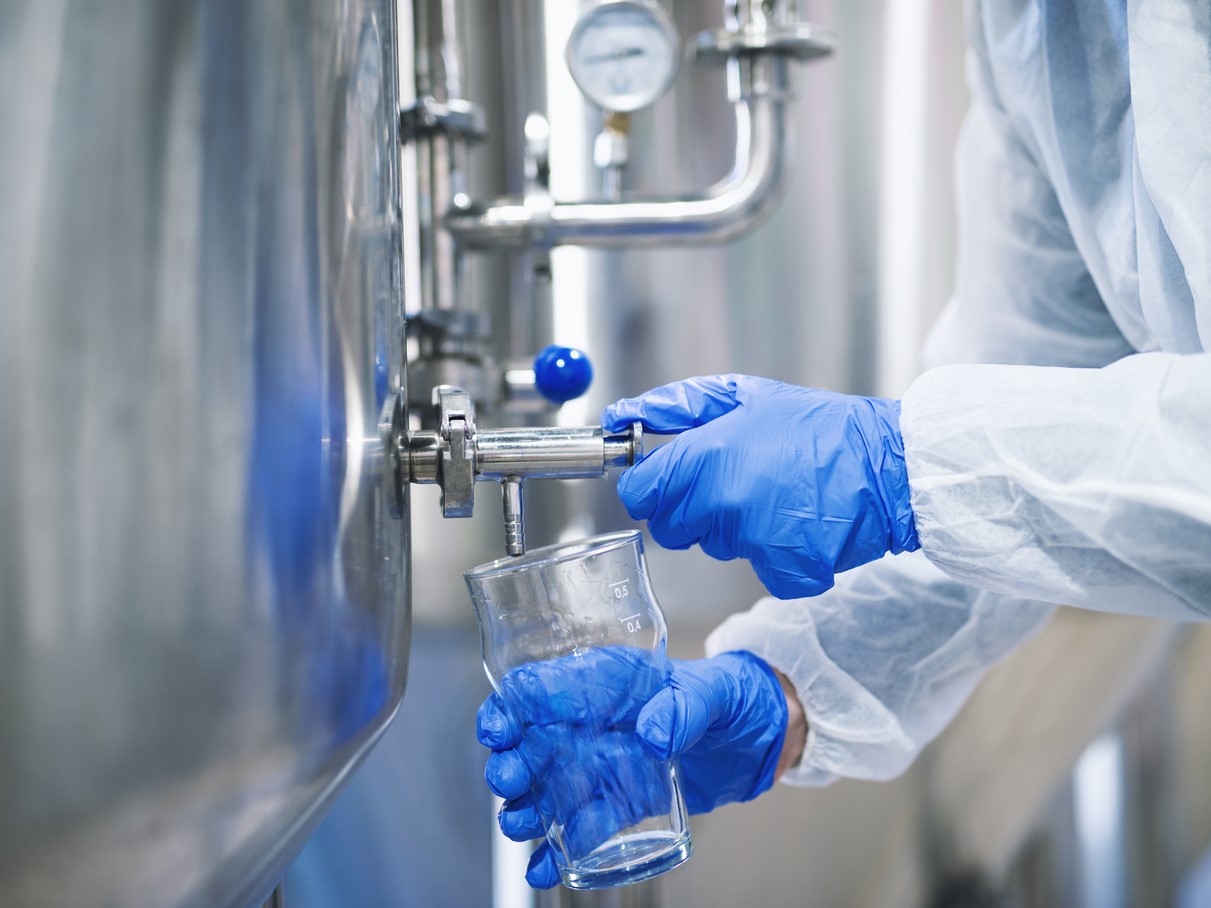 Close up view of hands in protective gloves opening tap valve and filling glass with samples from industrial machine reservoir.